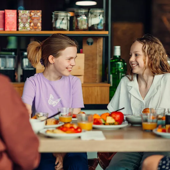 Kaksi lasta istuvat hymyillen ruokapöydän ääressä, lautasilla ruokaa ja lasit appelsiinimehua, taustalla hyllyt ja purkit.