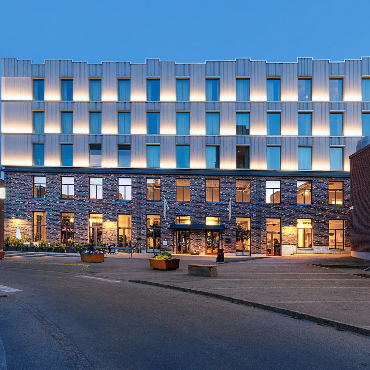 A modern building with a lit facade stands amidst an urban setting, surrounded by empty streets and additional brick structures. The sky appears early evening, with a clear blue hue.
