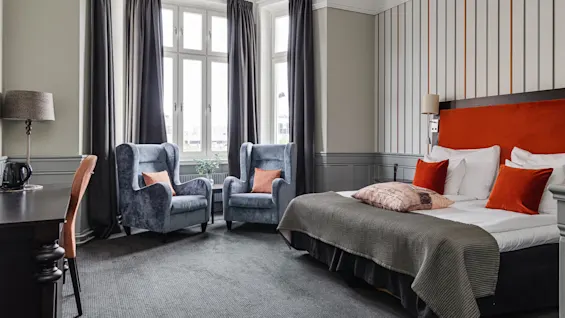 A bed with orange pillows rests against a striped wall, flanked by armchairs and a desk, in a sunlit room with large windows and heavy curtains.