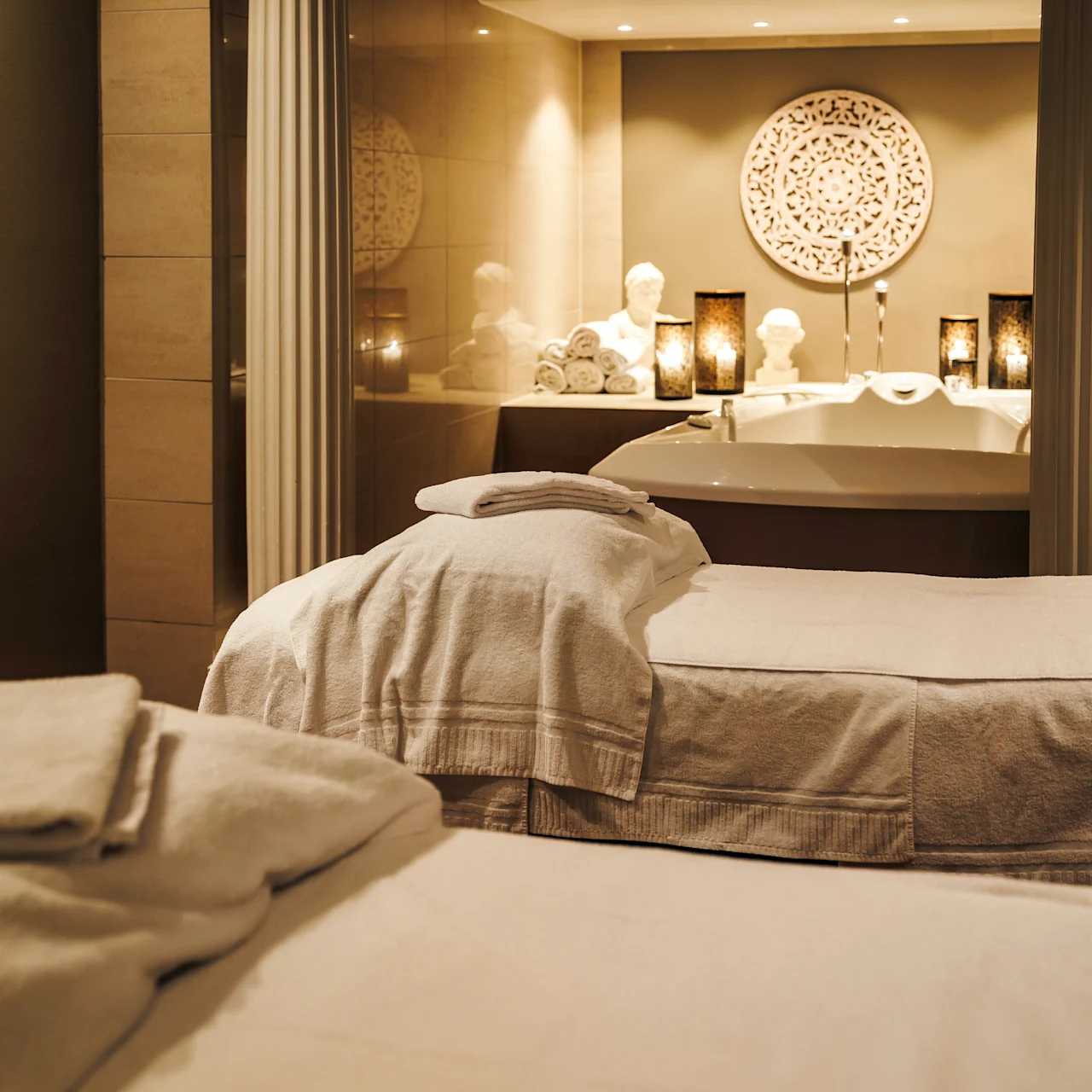Massage tables covered in white towels rest in a softly lit spa room, with decorative candles and rolled towels surrounding a luxurious bathtub.