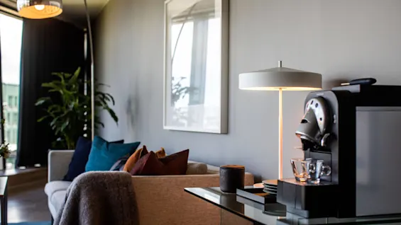 Kaffemaskin placerad på ett glasbord, bredvid en modern lampa, med en soffa dekorerad med färgglada kuddar i ett mysigt, välinredd vardagsrum med stora växter.