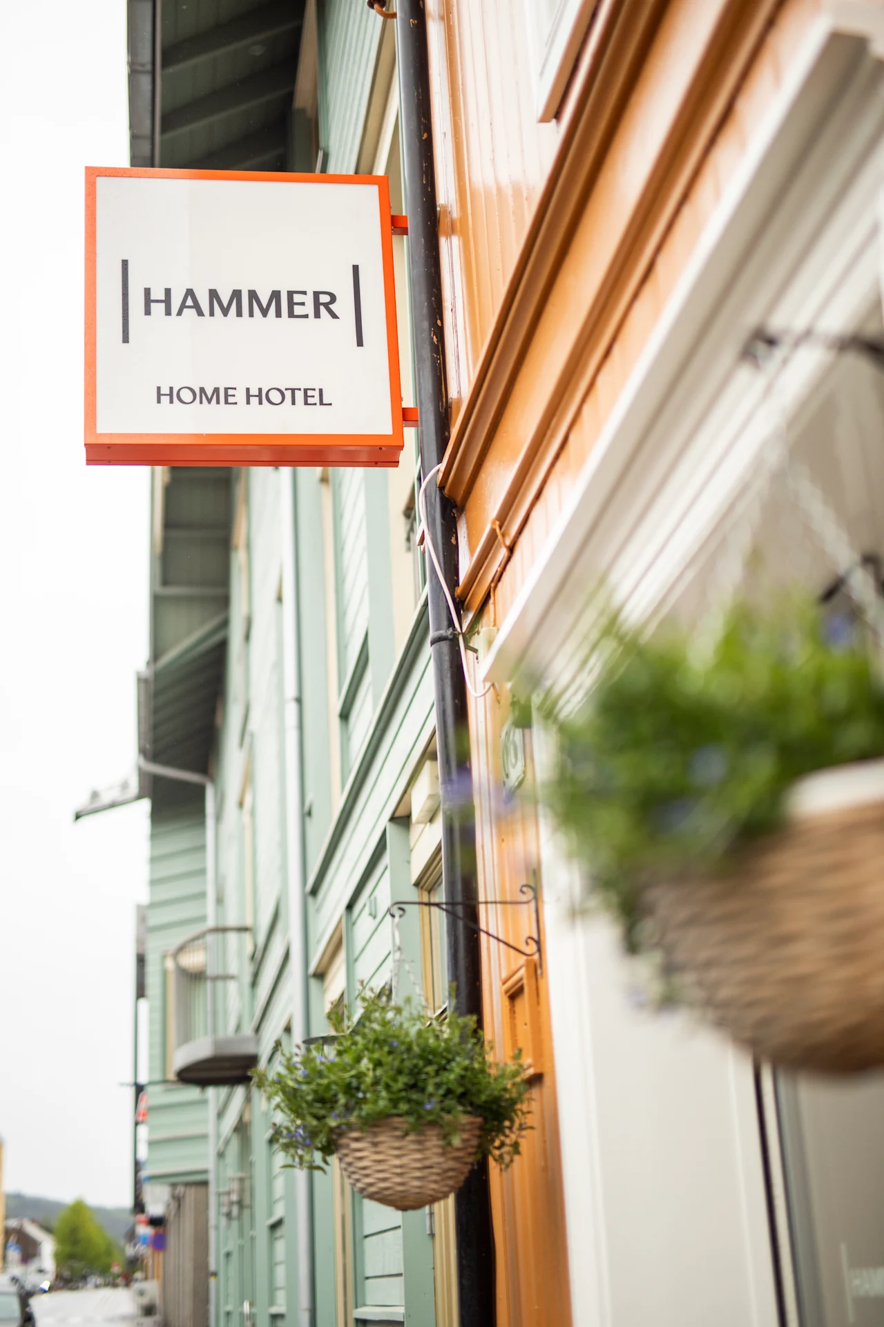 Skilt med teksten "HAMMER HOME HOTEL" henger på en fargerik bygningsfasade. To hengende kurver med grønt pryder forgrunnen. Gaten strekker seg inn i avstanden under en overskyet himmel.