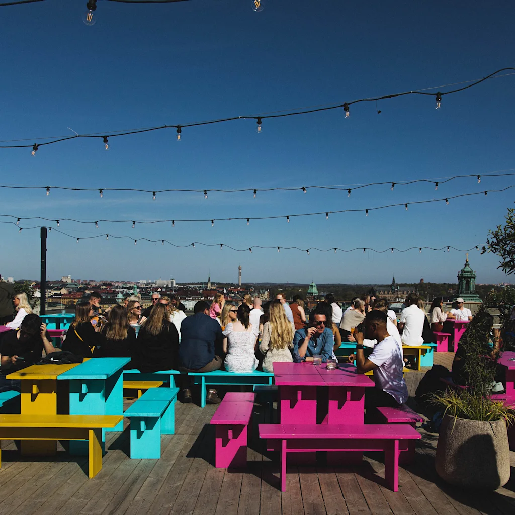 Folk nyder en solrig dag på Stockholm Under Stjärnorna (SUS) på At Six, en livlig tagbar med farverige borde.