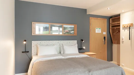 A cozy hotel room with a comfortable bed, bedside lamps, and framed landscape art. A wooden door with an emergency exit plan is visible.