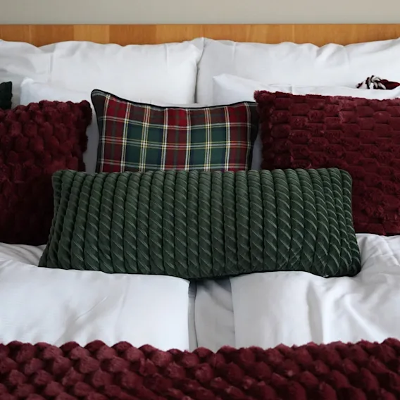 En bäddad hotellsäng med vita täcken och dekorativa kuddar i rött, grönt och rutigt mönster.