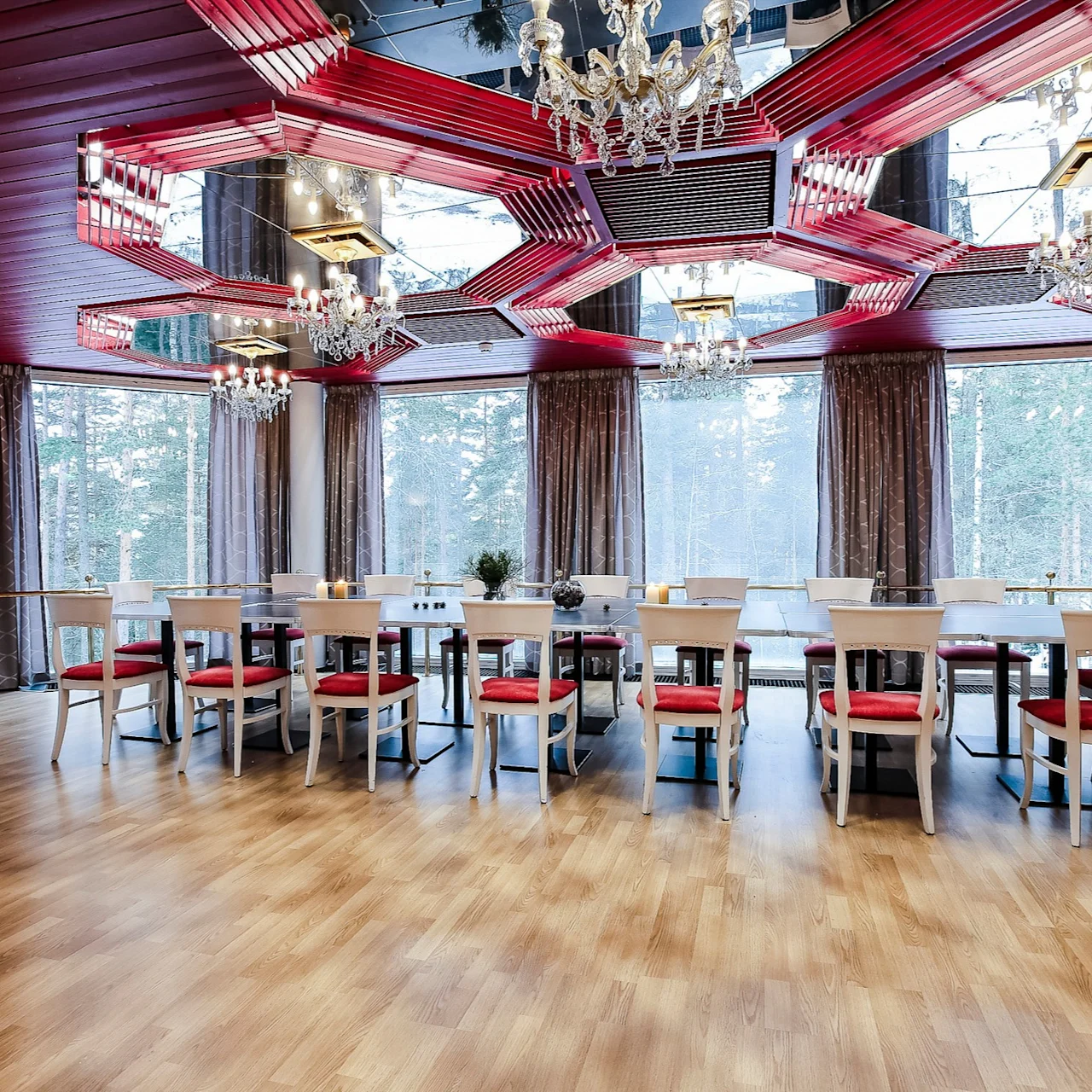 A long dining table with red-cushioned chairs is centered in a room with wooden floors and elaborate chandeliers. Large windows reveal a forested view outside.