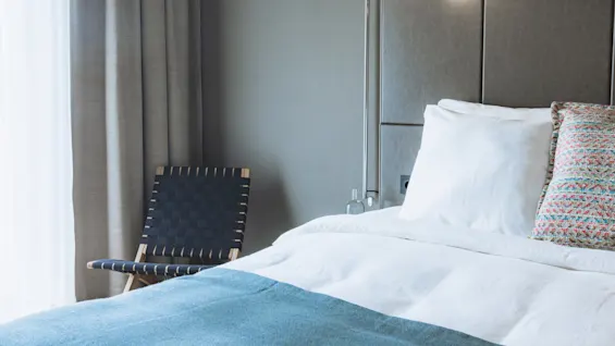 Seng med hvite og mønstrede puter, plassert ved siden av en moderne stol. En flaske og lampe pynter sengekanten, mens myke gardiner rammer inn vinduet.