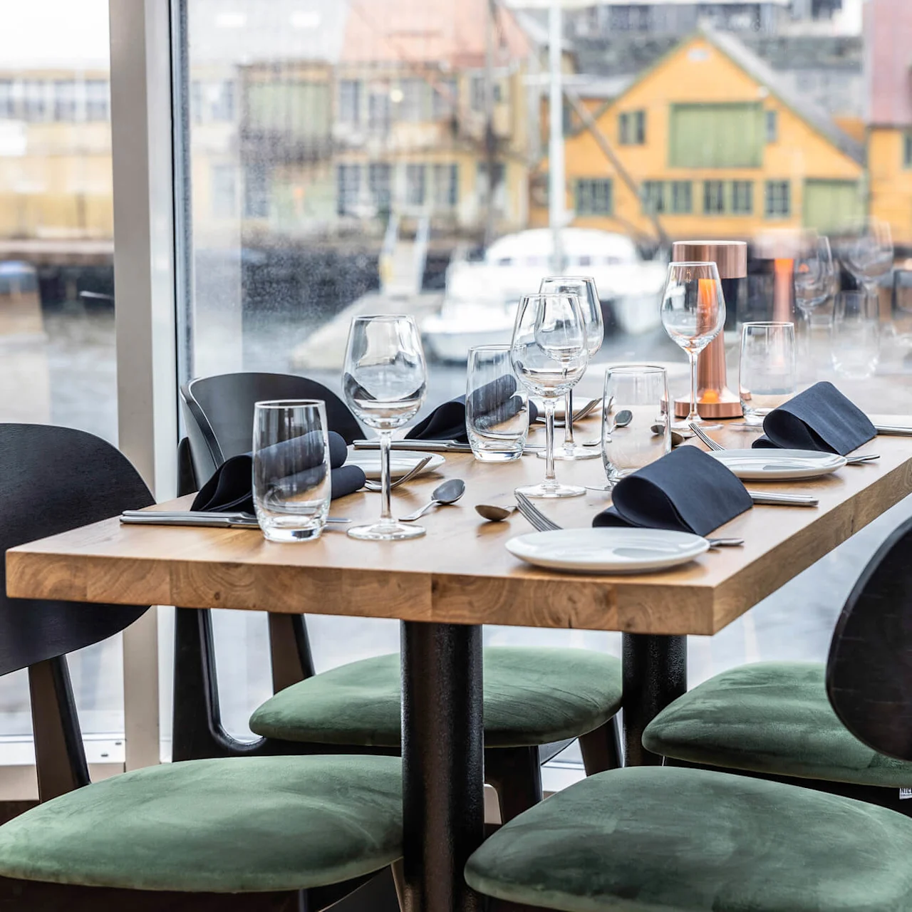 Ett träbord dukat med glas, tallrikar och vikta servetter; omgivet av moderna stolar med utsikt över en hamnmiljö genom stora fönster.