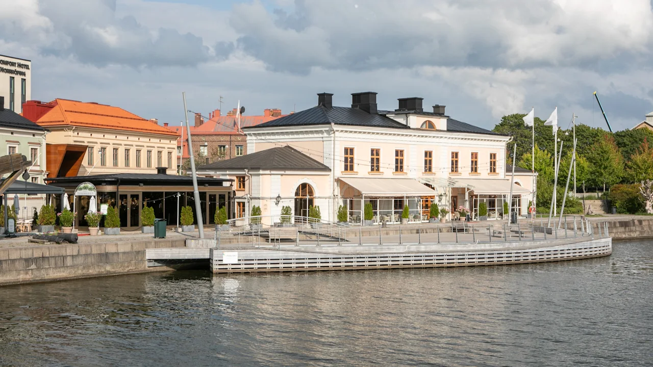 En bygning ved vandet med udeservering og skilt for HOTEL NORRKOPING i baggrunden, under delvist skyet himmel.