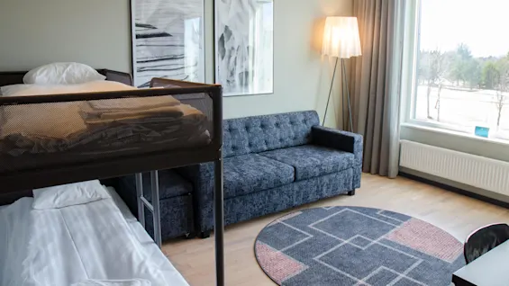 Våningssäng med sängkläder bredvid en mörk soffa. I närheten avger en golvlampa ljus. Två abstrakta målningar hänger på väggen. Ett stort fönster visar en utomhusscen.