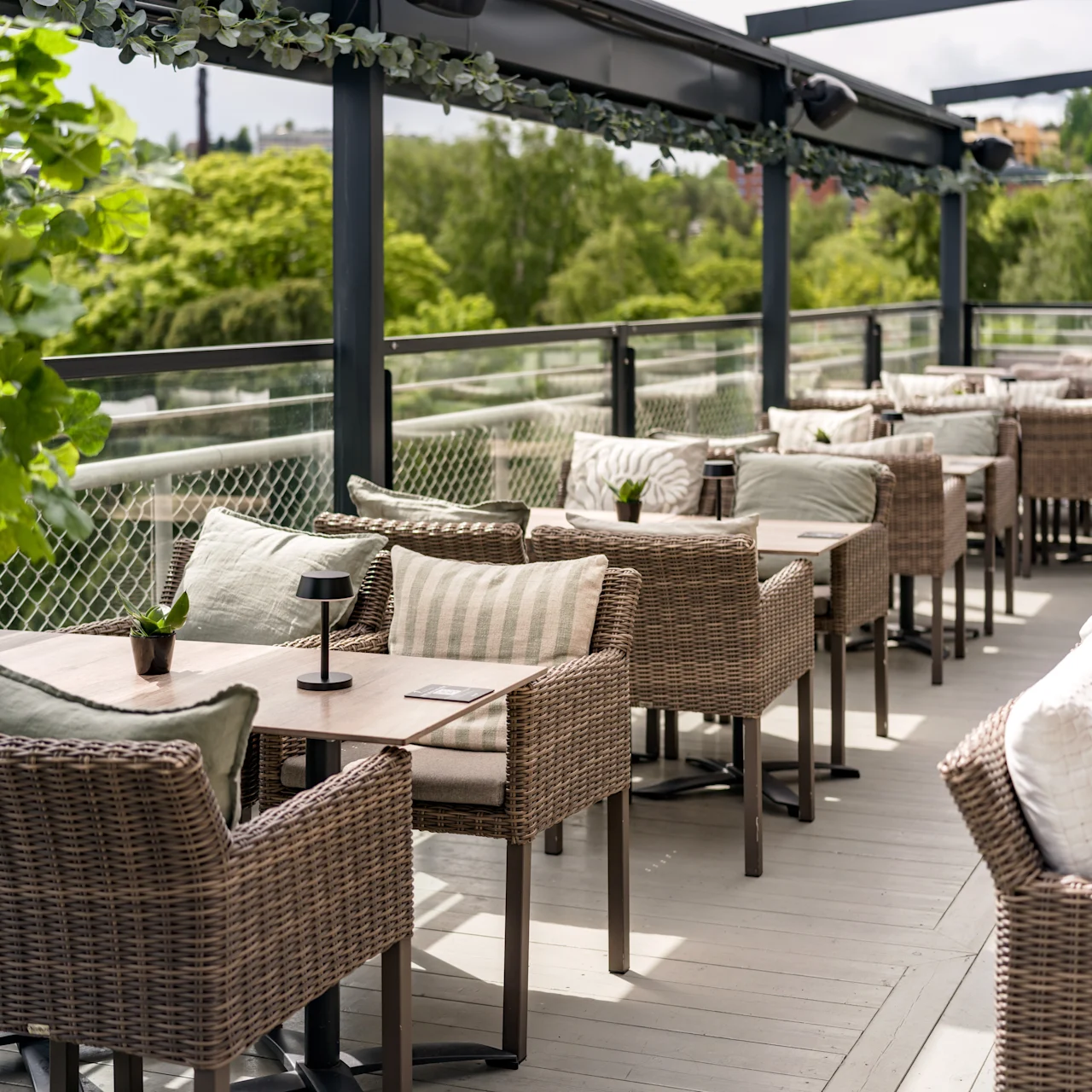 Outdoor dining area with wicker chairs and tables, surrounded by lush greenery. Overhead beams partially cover the space, providing shade on a sunny day. Small potted plants adorn each table.
