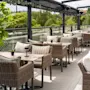 Outdoor dining area with wicker chairs and tables, surrounded by lush greenery. Overhead beams partially cover the space, providing shade on a sunny day. Small potted plants adorn each table.