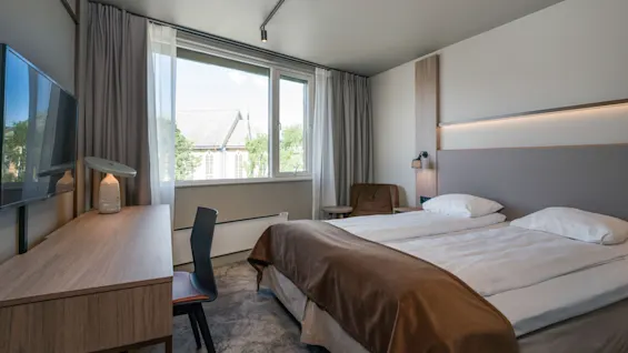En pænt redt dobbeltseng med hvidt sengetøj og et brunt tæppe står i et moderne hotelværelse. Et skrivebord med en lampe og stol er placeret ved vinduet.
