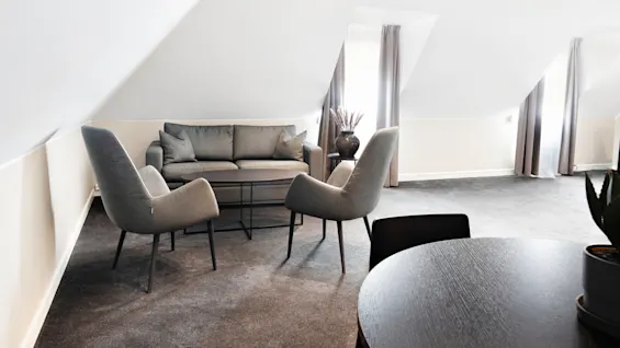 En moderne stue har en grå sofa og to lænestole omkring et sofabord på et gråt tæppe. Blødt naturligt lys strømmer ind gennem vinduer med gardiner.