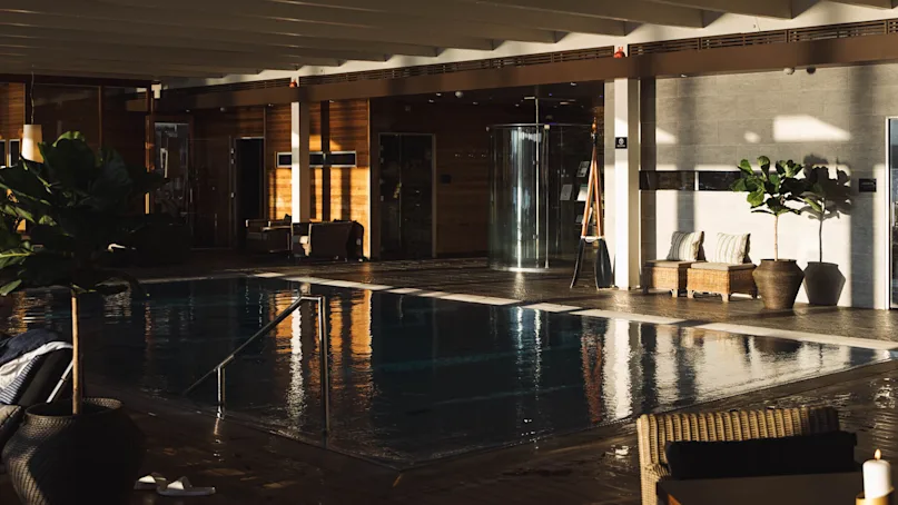 Swimming pool reflects sunlight, surrounded by lounge chairs and potted plants. Warm wooden panels and cushioned seating create a serene, indoor relaxation space.