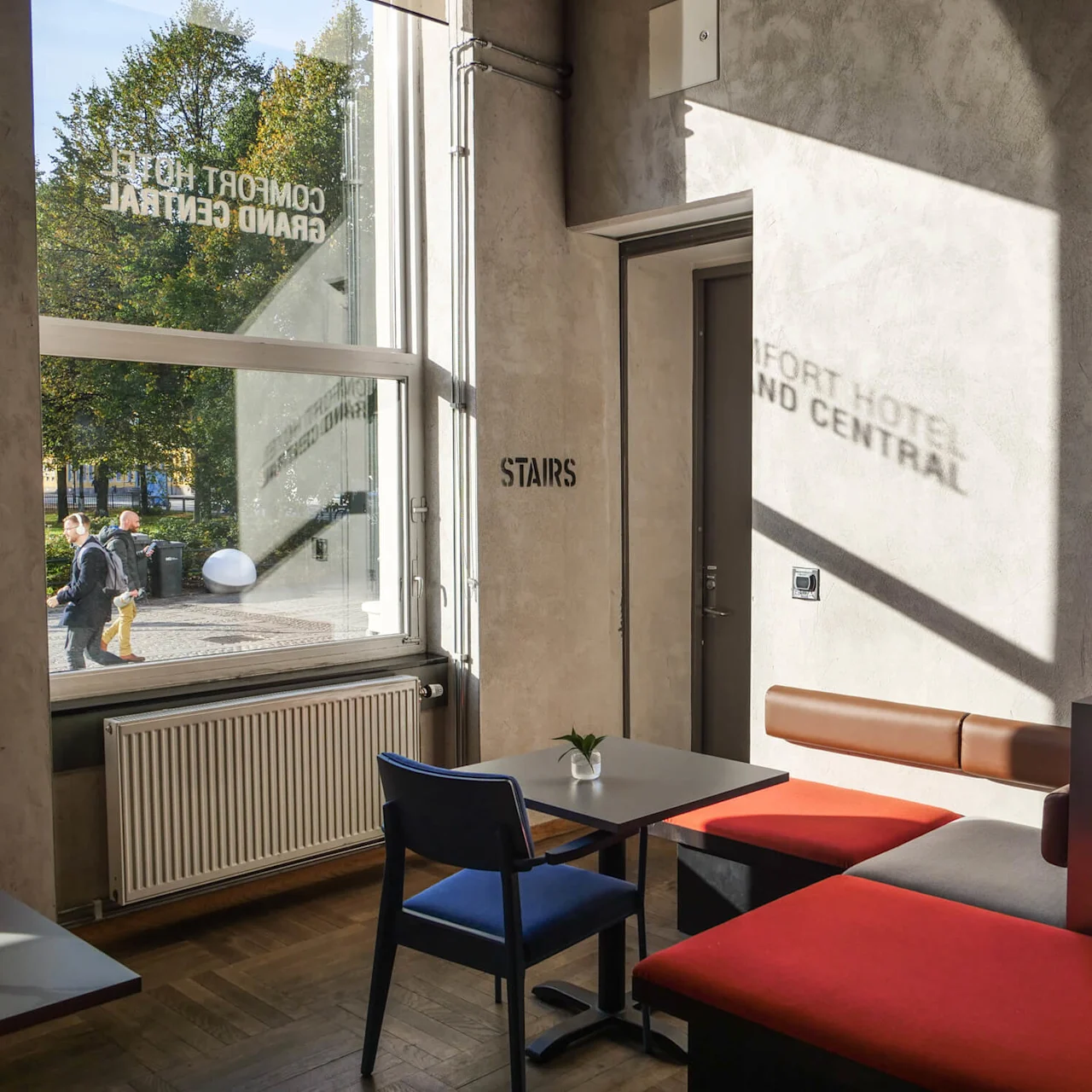 A window reflects "COMFORT HOTEL GRAND CENTRAL" and "STAIRS" onto a wall, illuminating a modern seating area inside. Outside, two people walk past trees in sunlight.