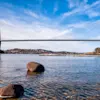 Hängbro sträcker sig över en bred flod, stödd av höga torn med kablar. Floden speglar den klara blåa himlen, omgiven av avlägsna kullar och utspridda träd längs stränderna.
