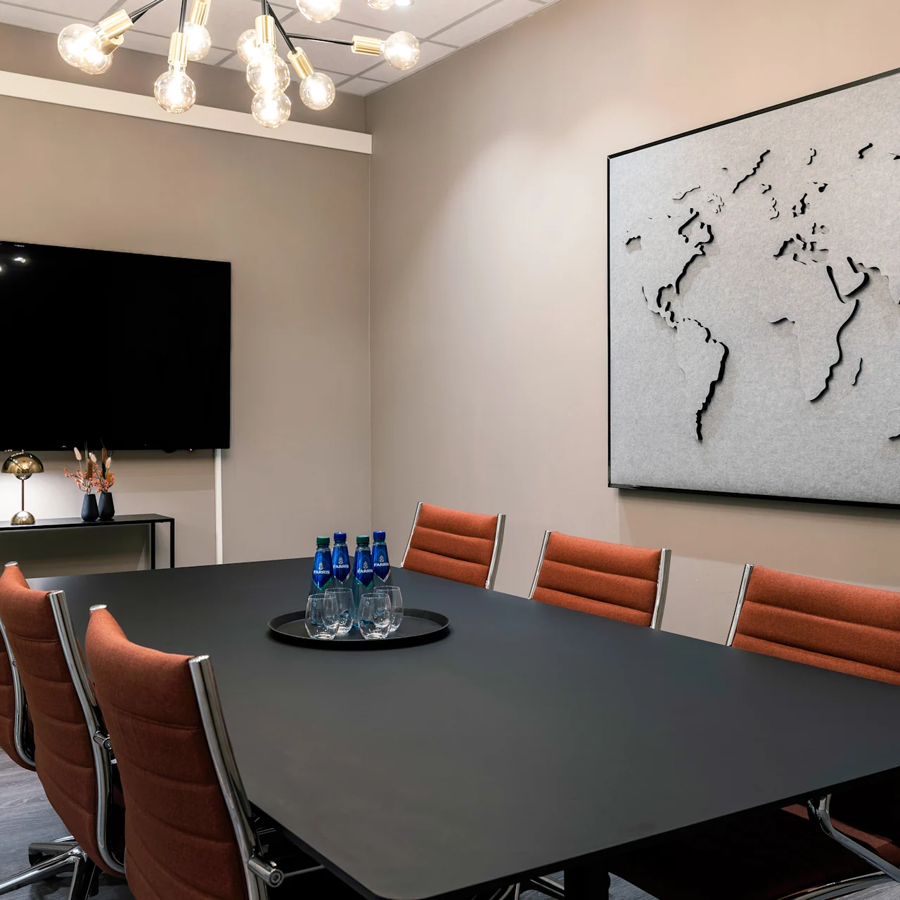Conference table with orange chairs holds a tray with blue water bottles and glasses; wall features a map art piece, next to a screen and decorative lamp.