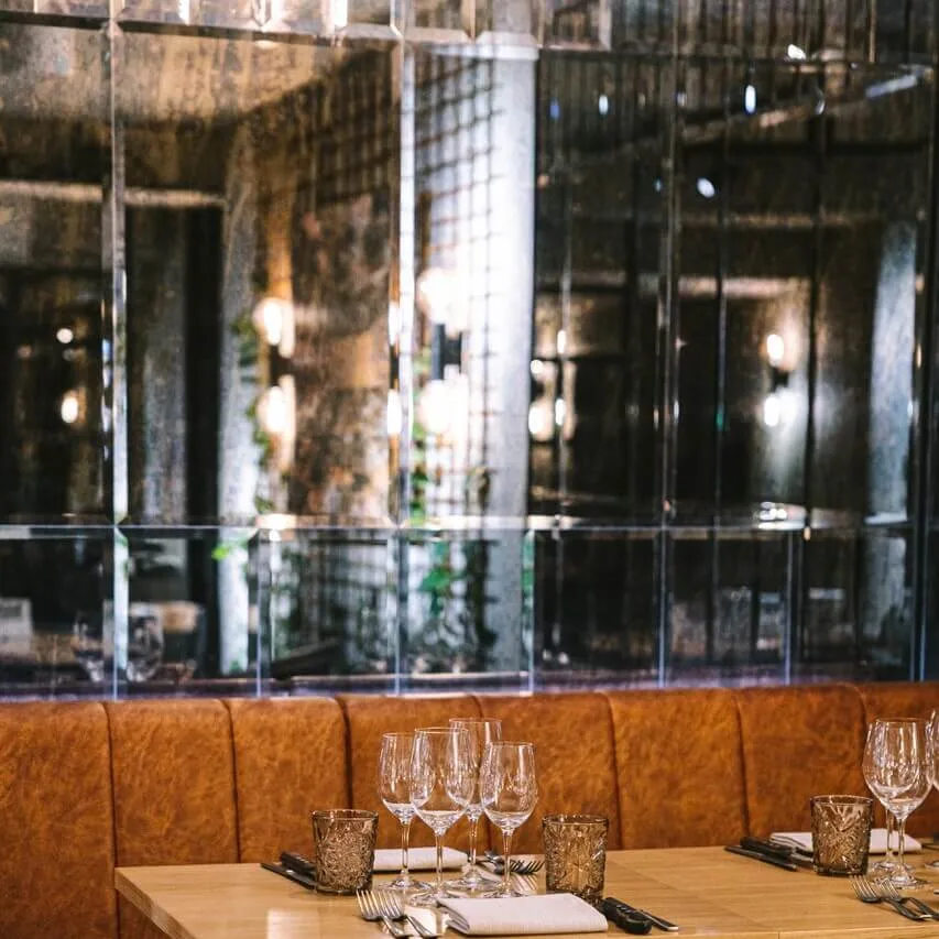 Et trebord er dekket med vinglass og bestikk på en brettet serviett. Det er omgitt av brune skinnseter, med reflekterende glassvegger i en svakt opplyst restaurant.