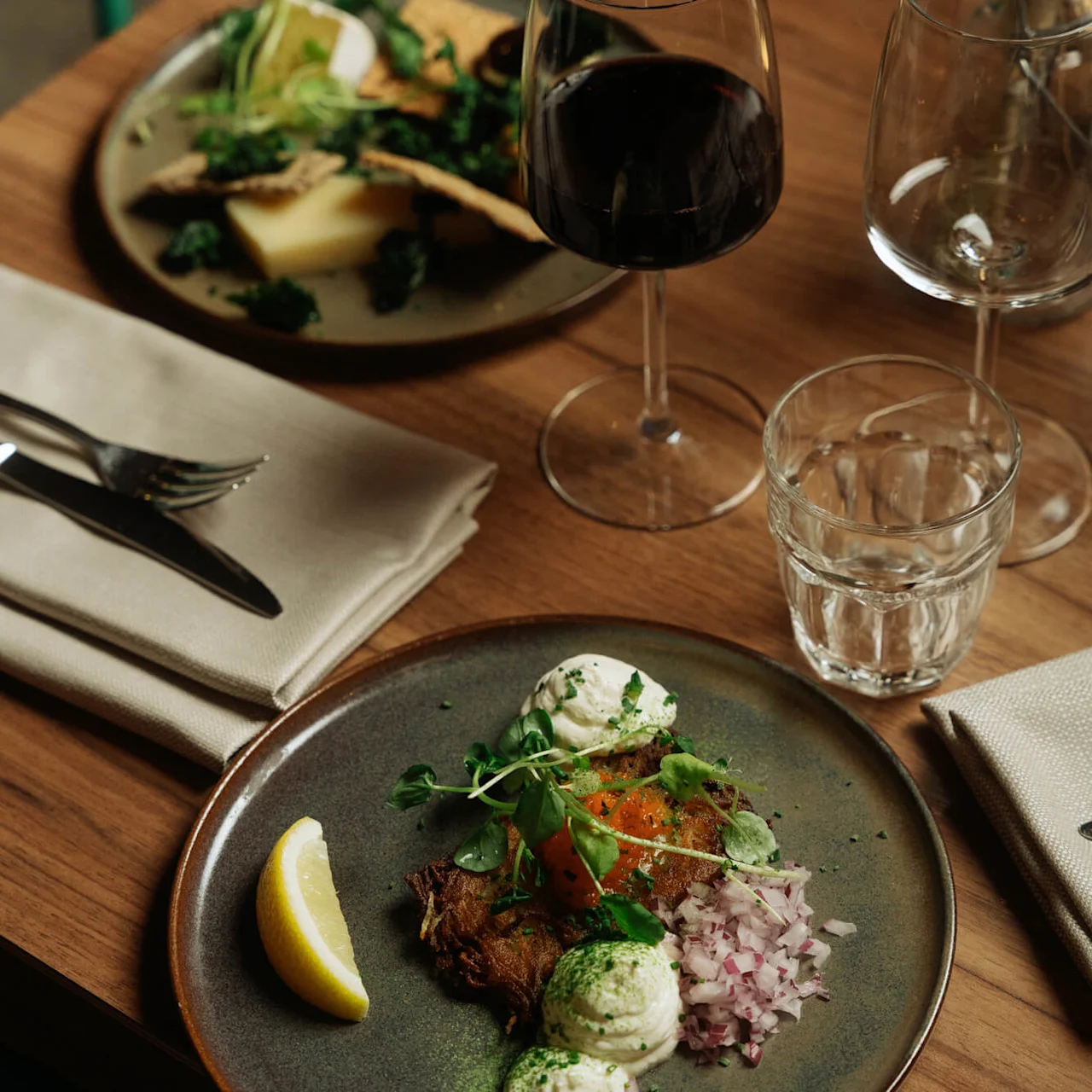 En tallrik med mat med en citronklyfta, garnerad med örter, står bredvid ett glas rött vin på ett träbord i en mysig matmiljö.