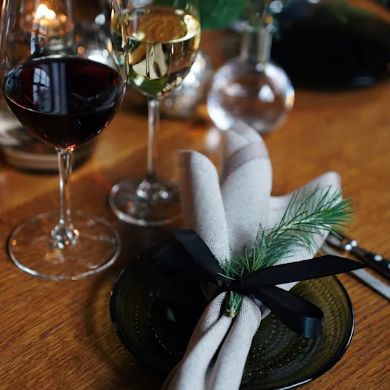 Festive dinner table setup with red and white wine glasses and a cloth napkin tied with a black ribbon and pine sprig on a green plate.