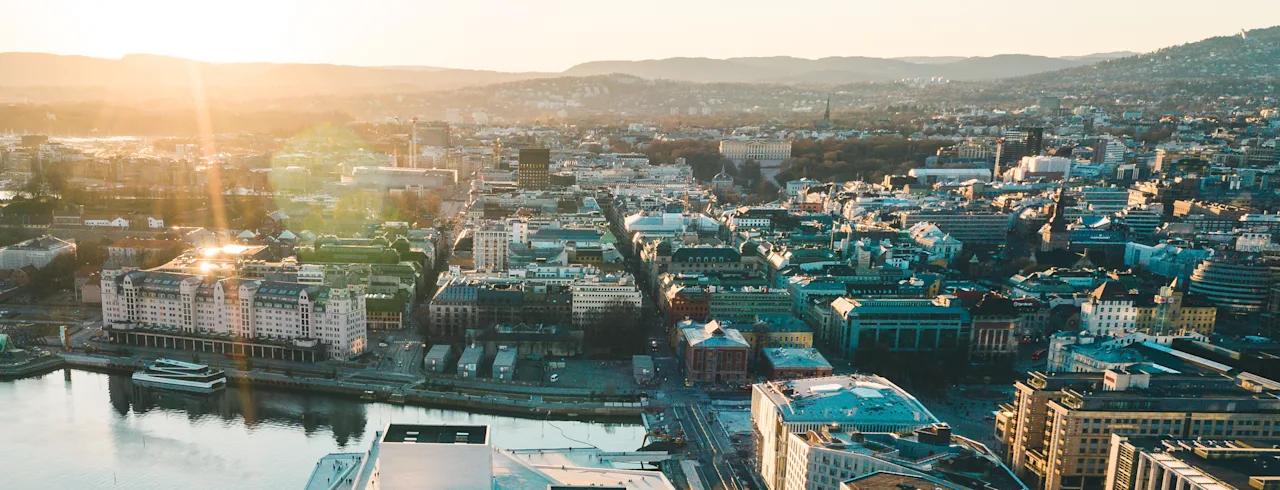 Aerial view of a city at sunset, with buildings, a waterfront, and mountains in the distance.
