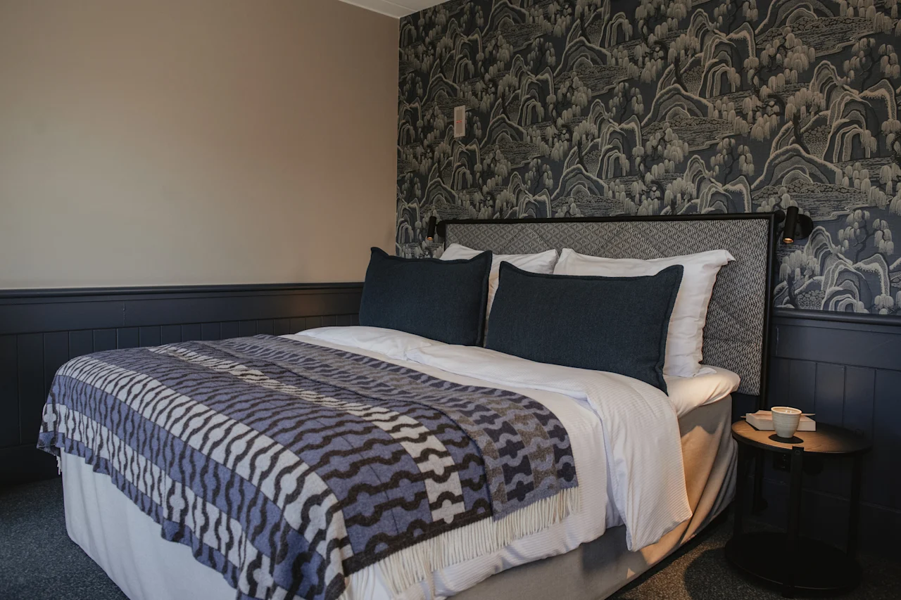 Ett mysigt sovrum med en säng med en mönstrad filt, mörka kuddar och en unik tapetdesign bakom sänggaveln.