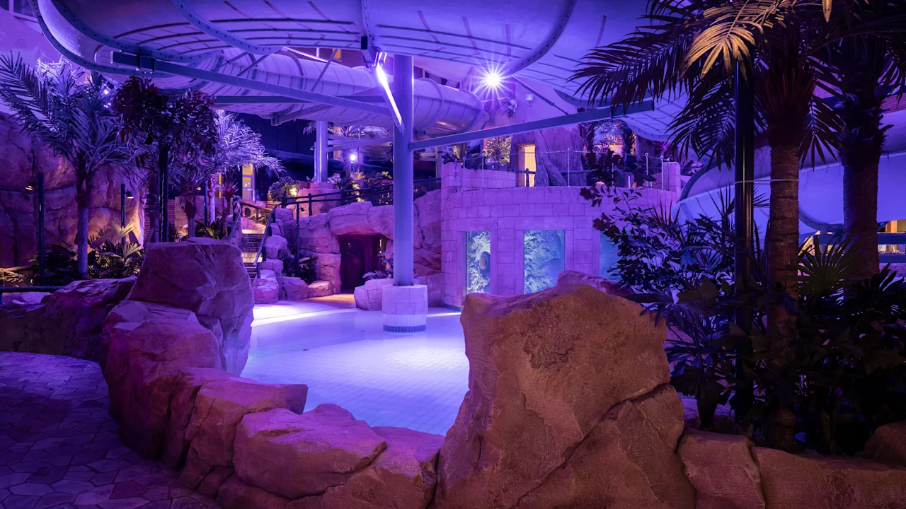 En indendørs vandland har en buet vandrutsjebane over en pool, omgivet af kunstige klippeformationer og tropiske planter, oplyst med lilla belysning.