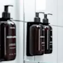 Lähikuva tummista pumppupulloista, joissa on BODY LOTION ja SHAMPOO, asennettuna valkoiselle kaakeloidulle kylpyhuoneen seinälle, heijastuen peilistä.