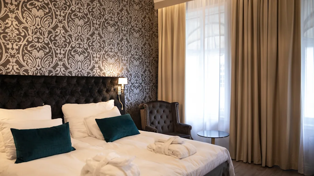 Et hyggeligt hotelværelse med en stor seng, elegant mønstret tapet og lyse vinduer med flagrende gardiner.