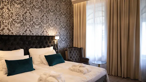 Et hyggeligt hotelværelse med en stor seng, elegant mønstret tapet og lyse vinduer med flagrende gardiner.