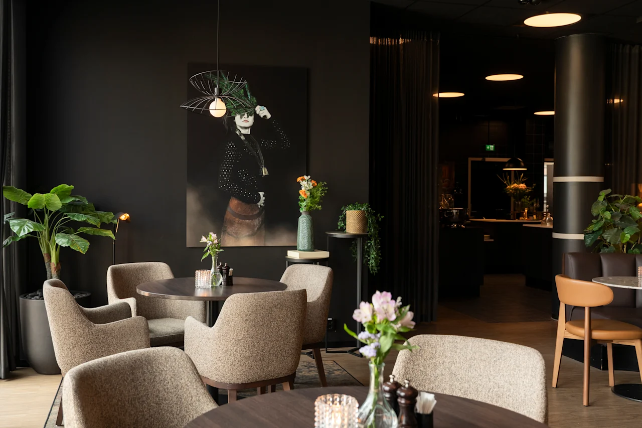 En stilig, dempet opplyst restaurant eller lounge med komfortable sitteplasser, planter og et stort kunstverk på en mørk vegg.