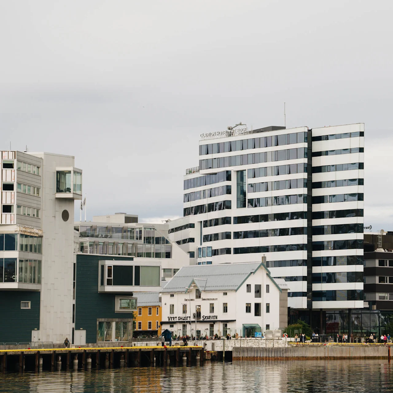 En høj, stribet bygning står ved vandet, med mindre strukturer i nærheden og skilte, der siger "VERT HUSET SKARVEN" og "CLARION HOTEL THE EDGE." Folk går langs dokken.