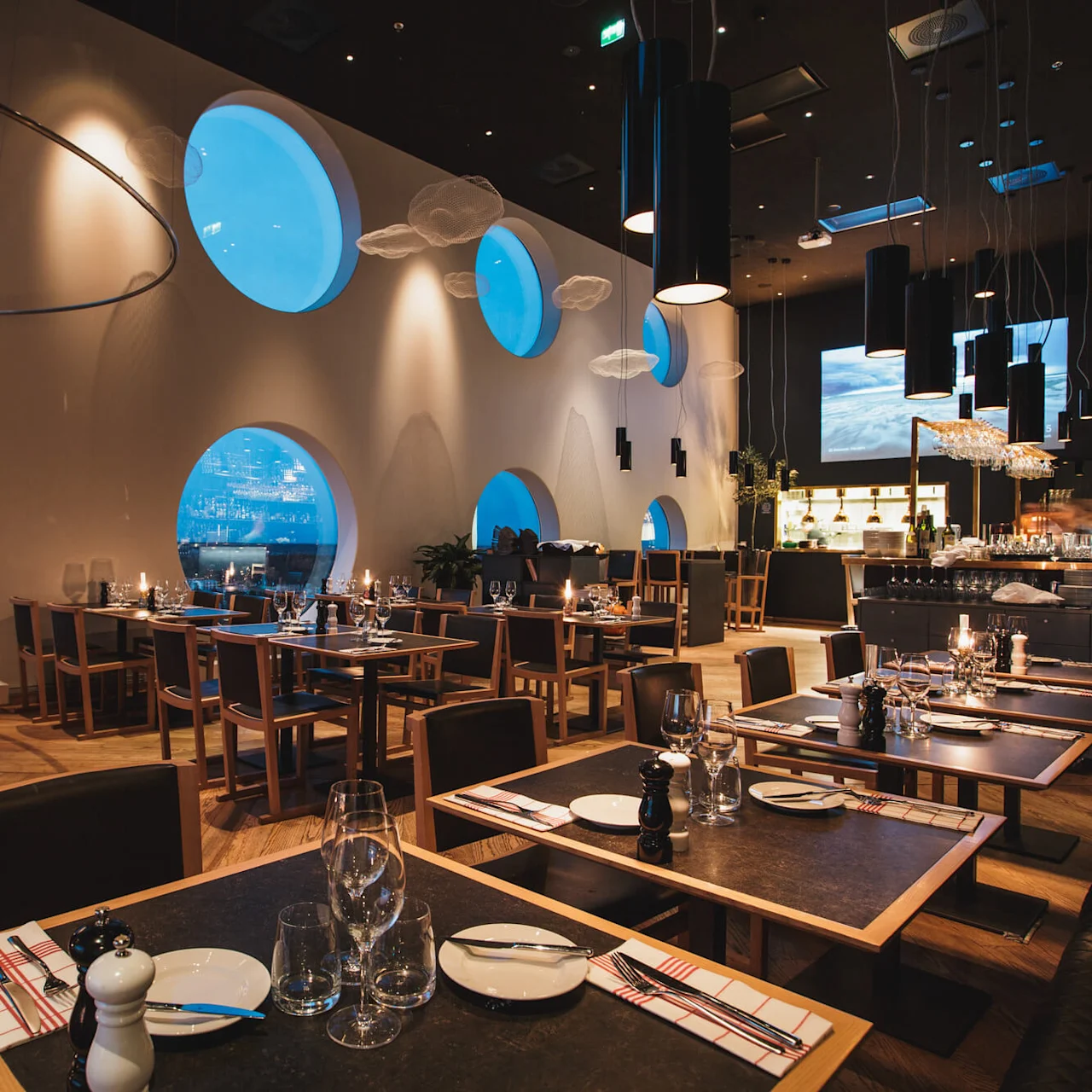 En moderne restaurant med runde vinduer med udsigt over byen, træborde dækket med glas og bestik og stemningsfuld belysning fra unikke lysekroner, der skaber en elegant spiseoplevelse.