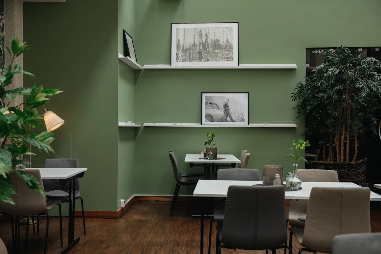 Café med trägolv och gröna väggar, med vita hyllor som visar svartvita inramade bilder. Bord och stolar är ordnade prydligt, omgivna av löviga växter, vilket skapar en mysig atmosfär.