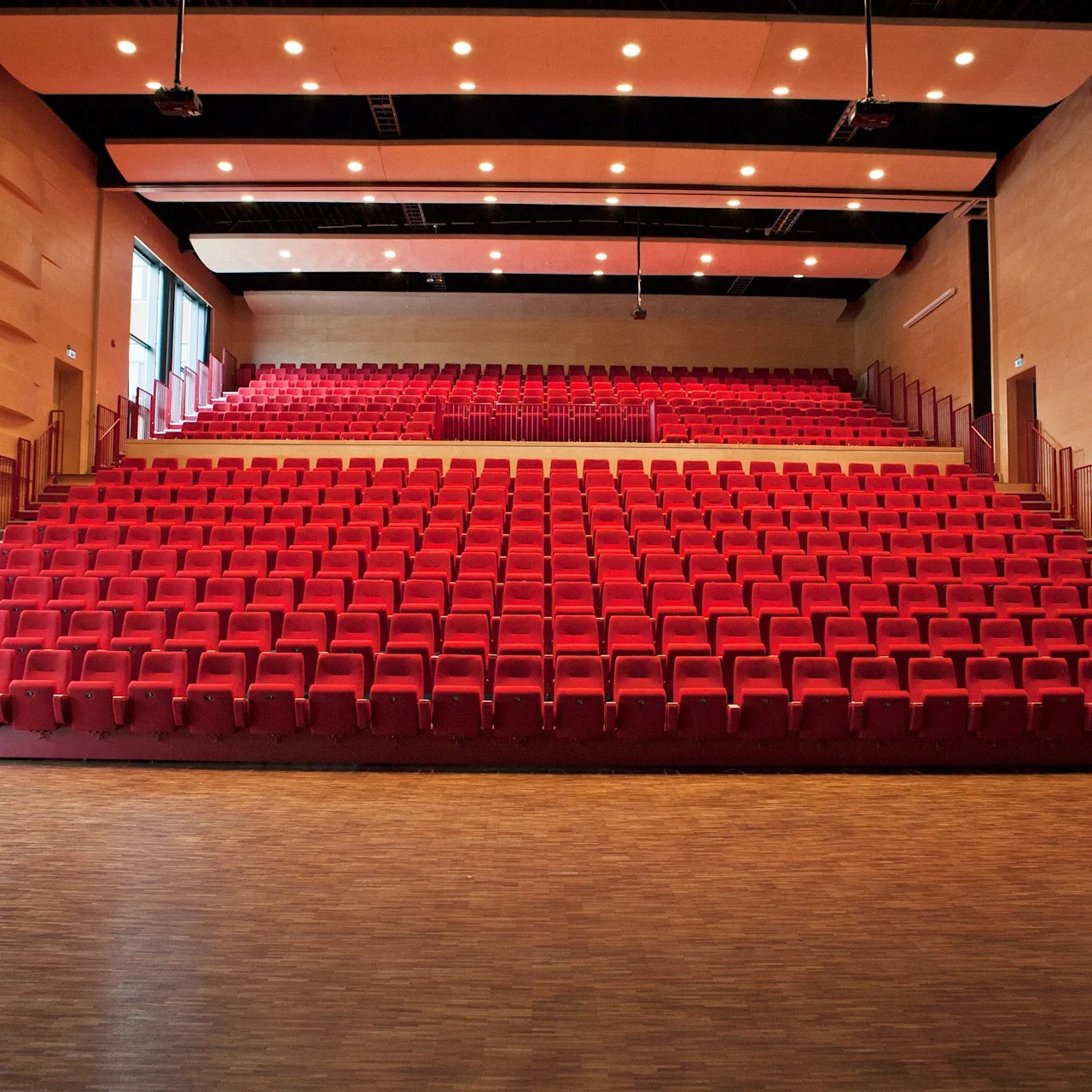 Rader med tomme røde teaterseter er arrangert i et trinnvis auditorium, vendt fremover. Rommet har tregulv og vegger, med taklys som lyser opp rommet.
