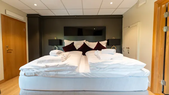 Et koselig hotellrom med en stor seng, hvitt sengetøy og mørkerøde puter. Nattbordslamper og tredører rammer inn det innbydende rommet.