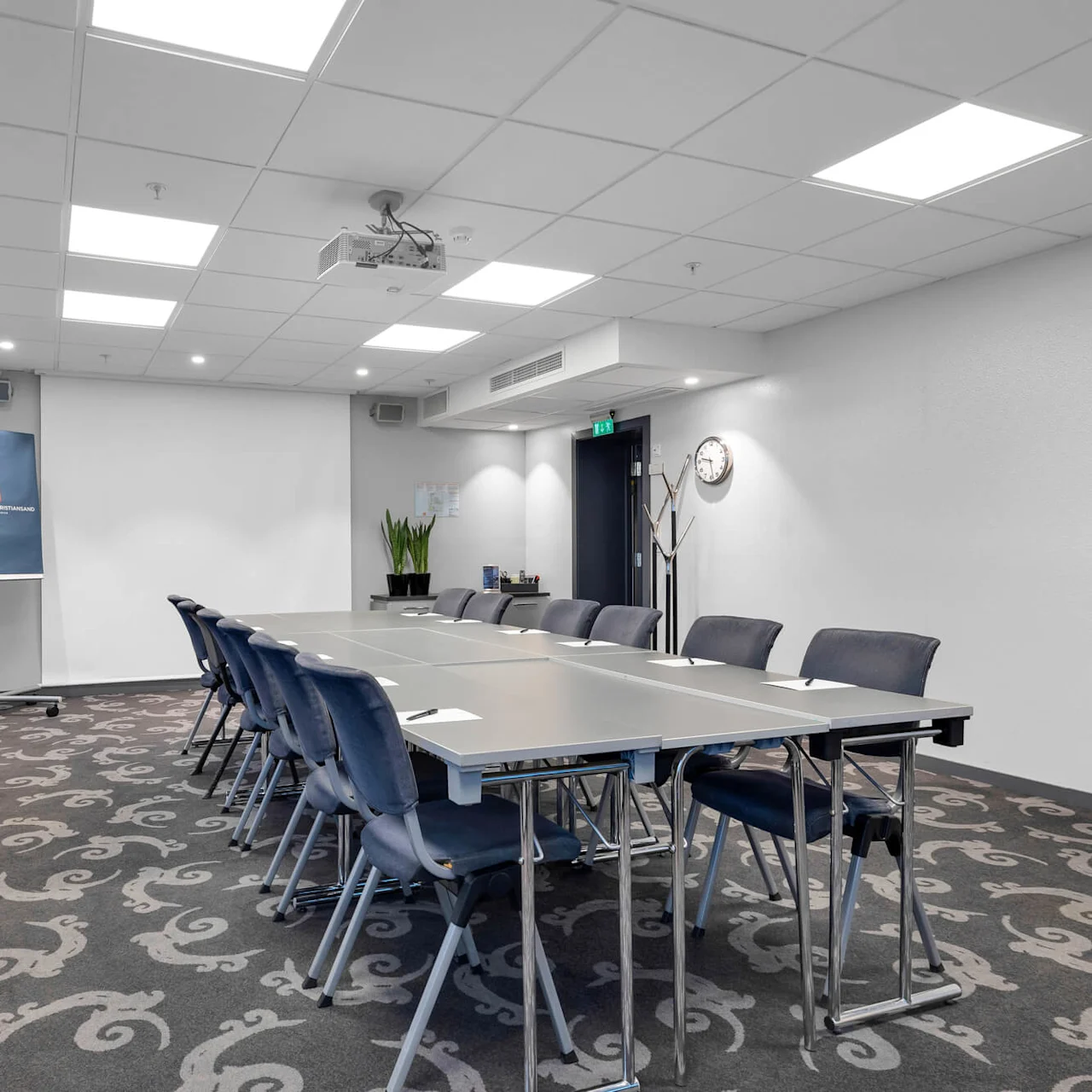 Et rektangulært konferencebord er omgivet af blå stole i et tæppebelagt møderum. Et whiteboard med teksten "CLARION HOTEL TREND STANDARD" står ved gardiner.