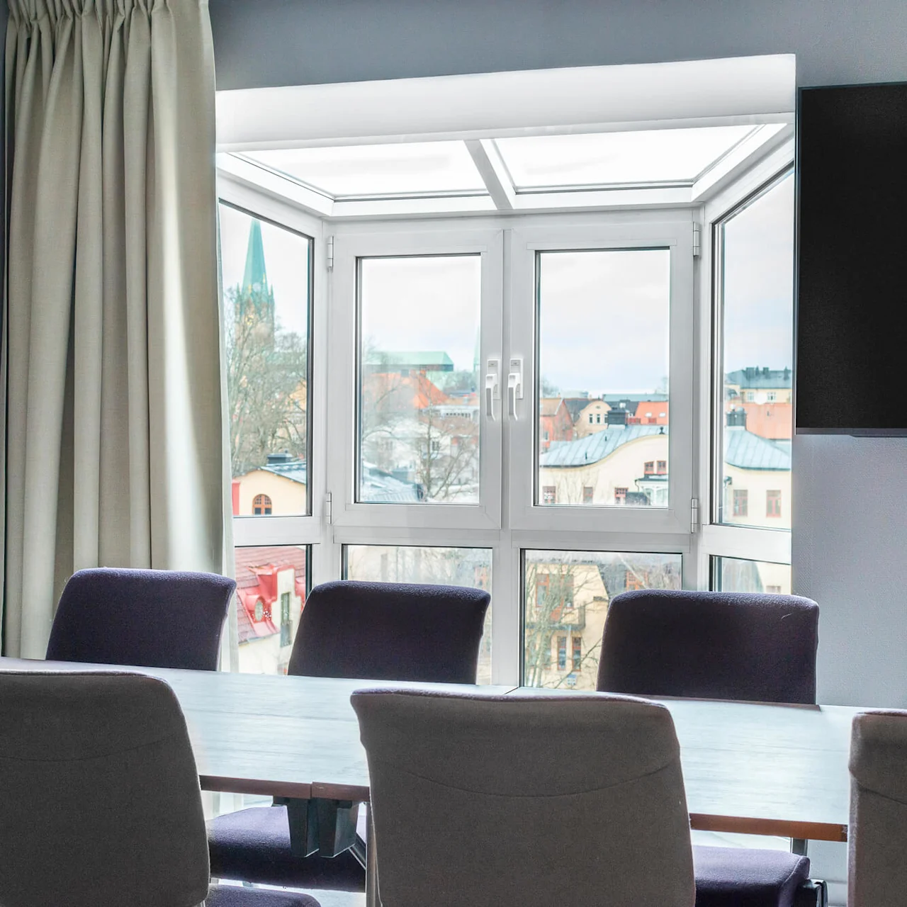 Ett konferensbord omges av stolar framför ett stort fönster med utsikt över tak och en avlägsen kyrkspira. En gardin och en platt-TV omger scenen.
