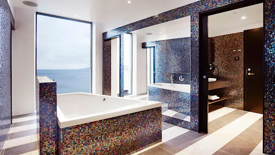Et luksuriøst bad med et stort badekar med flerfargede mosaikkfliser ved siden av et vindu med utsikt over havet under en overskyet himmel. Den moderne innredningen er minimalistisk og elegant.