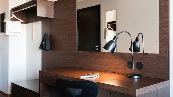 Ett skrivbord i trä har en anteckningsbok och en penna, belyst av en svart skrivbordslampa i ett modernt hotellrum. En spegel reflekterar en garderob med galgar och en tvättpåse.