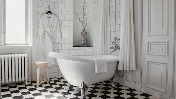 Klofotbadkar står med en handduk draperad över kanten; vit morgonrock hänger i närheten. Svartvitt schackbrädsgolv med kaklade väggar omger det. Radiator och pall ger detaljer.