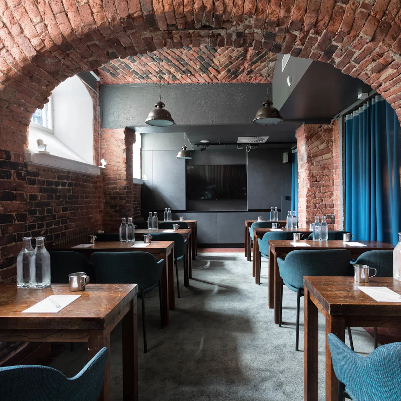 A conference room restaurant interior with wooden tables, blue upholstered chairs, and industrial pendant lights, providing a cozy, inviting atmosphere.