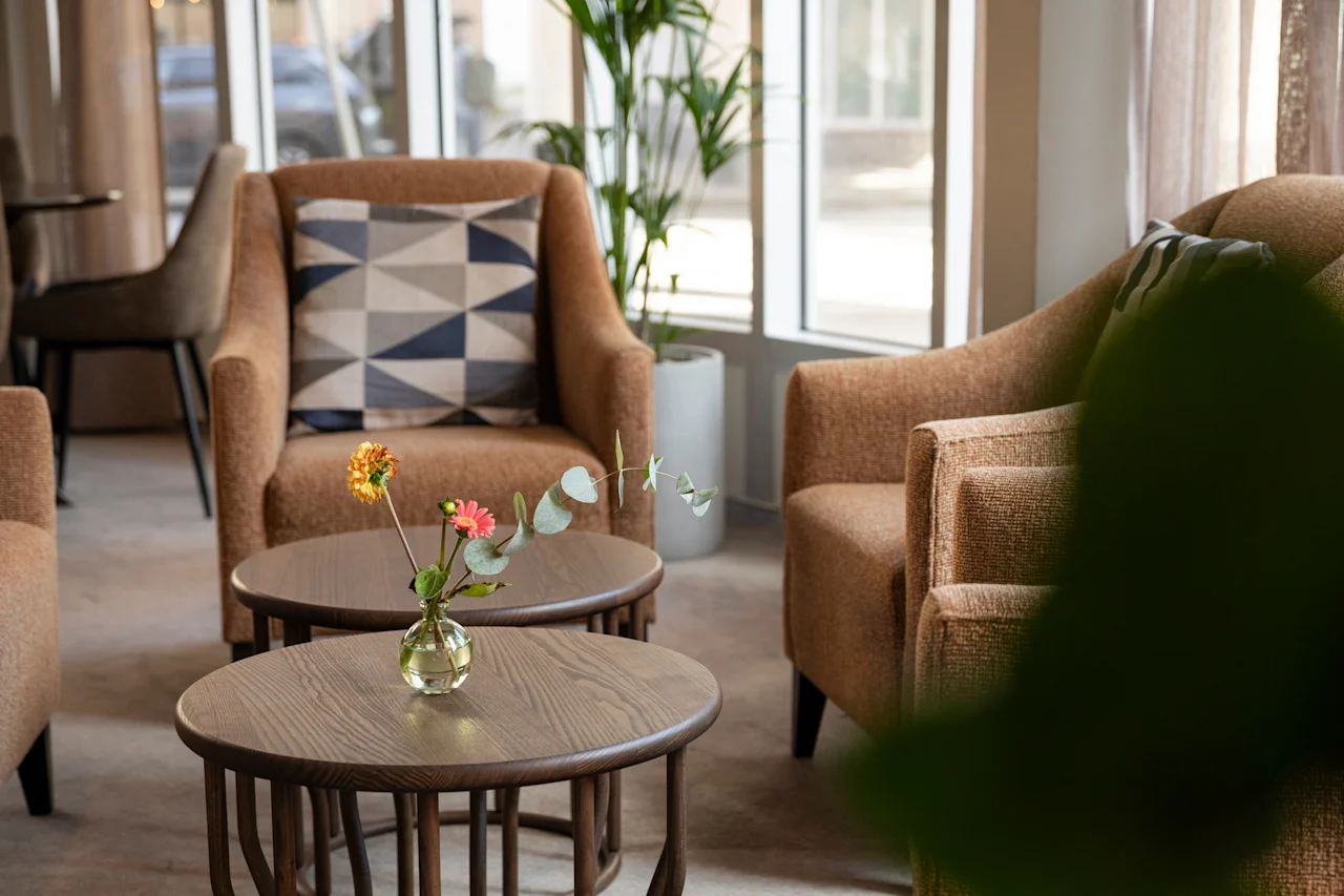 En mysig hotellounge med två fåtöljer, runda träbord och en vas med blommor, badad i naturligt ljus.