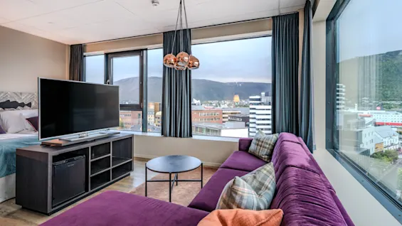 Et moderne hotellrom med en lilla seksjonssofa, en stor TV og store vinduer med utsikt over en by og fjell.