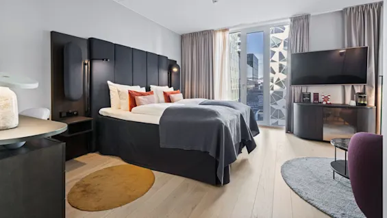 Modern bed with gray bedding, accented by orange pillows, placed against a dark headboard. Situated in a spacious room with a wall-mounted TV, large windows, and light gray curtains.