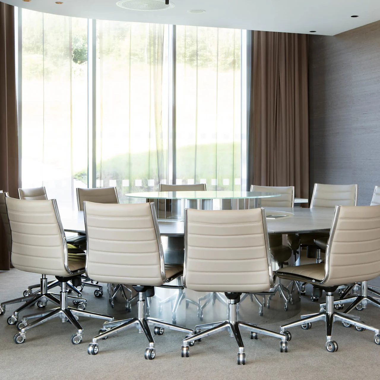 A round table surrounded by beige office chairs, situated in a modern conference room with a large window and a wall-mounted flat-screen TV.