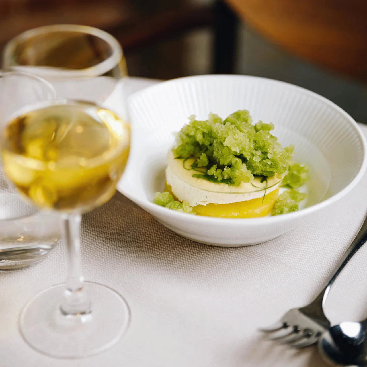 A dessert topped with green granita sits in a white bowl on a dining table, accompanied by a glass of white wine and cutlery nearby.