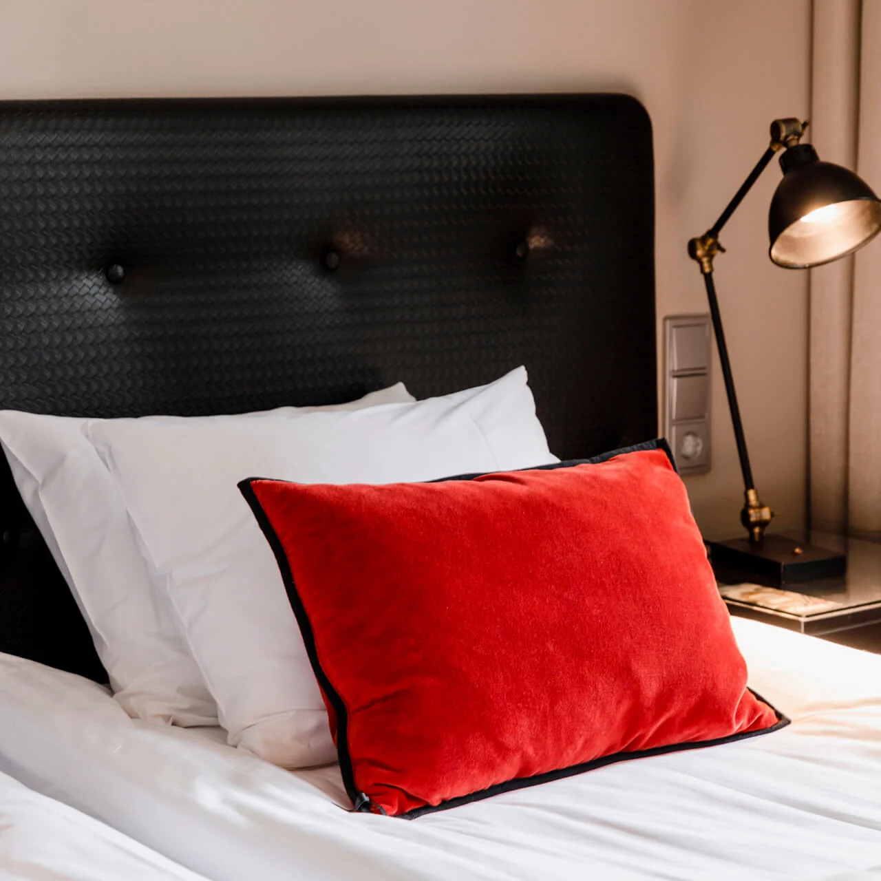 Seng med en rød pute hviler på hvite laken, ledsaget av en svart lampe på et sidebord, omgitt av beige gardiner.