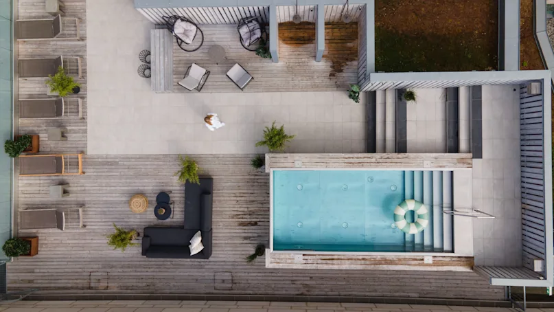 A rooftop terrace features a rectangular pool, lounge chairs, and seating areas, with a person walking across the wooden deck. Potted plants decorate the space.